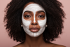 “High-end beauty photo of a dark-skinned Black woman wearing a white clay face mask in geometric strokes, standing against a blush-pink background. Her curls frame her face and she gazes at the camera with a serene confident expression. Lighting is soft and even, emphasizing contrast between mask and skin. Luxury editorial skincare aesthetic.”