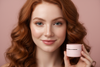 Ultra-realistic beauty portrait of an Irish woman with ginger curls, grey eyes, and faint freckles smiling softly toward the camera. Background is a pastel rose-pink. Skin is luminous and natural. She holds a pastel-pink OliviaSkin.co cream jar in one hand. Lighting is premium soft studio lighting.”